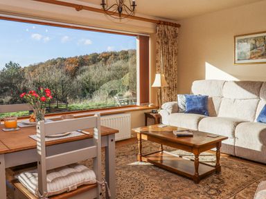 A living room with a sofa, coffee table, dining table with chairs, and a large window showing an outdoor view at Sherway Dairy in Clyst Hydon near Bradninch