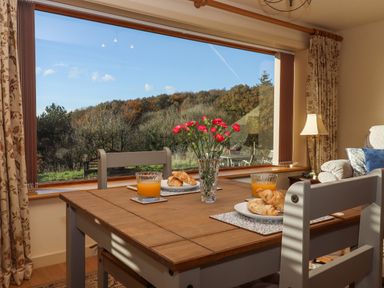 A dining area with a wooden table set with croissants and orange juice next to a large window showing trees outside at Sherway Dairy in Clyst Hydon near Bradninch