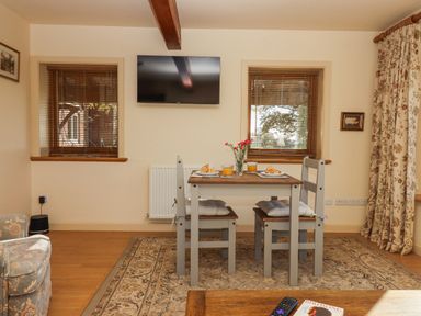 A dining area with a wooden table and two chairs with cushions two plates with croissants two glasses of orange juice and a vase with flowers two windows with blinds a television mounted on the wall and a patterned rug on the floor at Sherway Dairy in Clyst Hydon near Bradninch