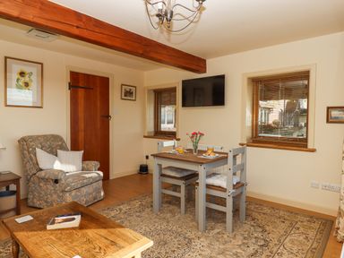 A dining table with two chairs and cushions a floral armchair a wooden coffee table with a book a wooden door two windows with blinds and a wall-mounted television in a living room at Sherway Dairy in Clyst Hydon near Bradninch