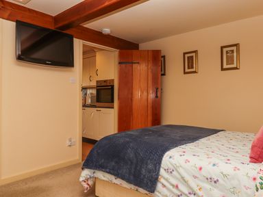 A bedroom with a bed covered with a floral duvet and navy blanket a door open to a kitchen with cream cabinets and an oven a wall-mounted TV two framed pictures and wooden ceiling beams at Sherway Dairy in Clyst Hydon near Bradninch