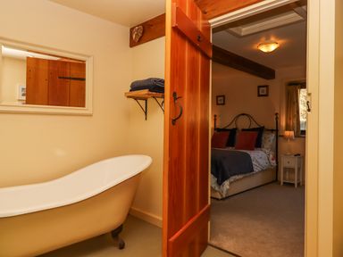 A bathroom with a clawfoot bathtub and a wooden door open to a bedroom with a bed and bedside table at Sherway Dairy in Clyst Hydon near Bradninch