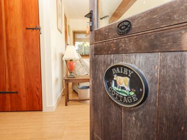 A wooden door with a dairy cottage sign partially open to a room with a side table lamp and armchair at Sherway Dairy in Clyst Hydon near Bradninch