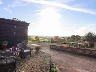 An outdoor patio with wicker chairs and a table next to a dark wooden building and a seating area with a wooden table and chairs surrounded by plants and trees with hills in the background at Sherway Dairy in Clyst Hydon near Bradninch