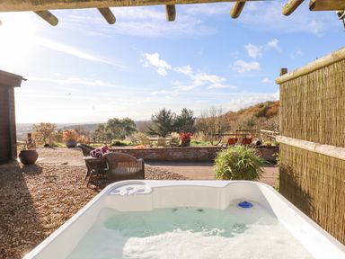 An outdoor hot tub with wicker chairs and a table on gravel beside a garden area with trees and hills at Sherway Dairy in Clyst Hydon near Bradninch