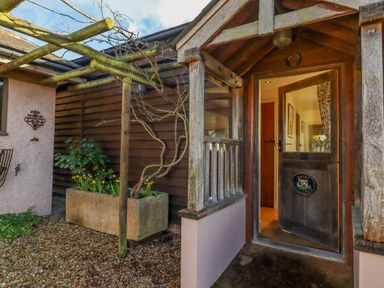 An exterior entrance with a wooden door opening to a hallway and a planter with plants beside a wooden pergola at Sherway Dairy in Clyst Hydon near Bradninch