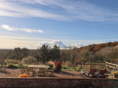 An outdoor patio area with wooden table and chairs and two lounge chairs overlooking trees and hills at Sherway Dairy in Clyst Hydon near Bradninch