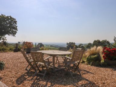 An outdoor patio with a round wooden table and six wooden chairs surrounded by plants and a view of distant hills at Sherway Dairy in Clyst Hydon near Bradninch