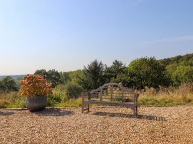 A wooden bench and a pot with flowers on a gravel area with trees in the background at Sherway Dairy in Clyst Hydon near Bradninch