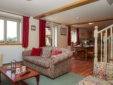 A living room with patterned sofas red cushions red curtains wooden dining table with chairs and kitchen area at Sherway Shippen in Clyst Hydon near Bradninch