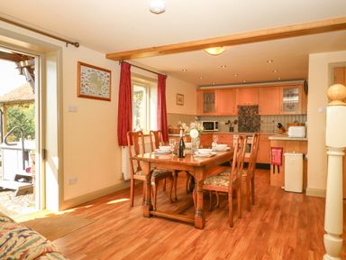 A dining area with wooden table and chairs set for four next to a kitchen with wooden cabinets and tiled backsplash at Sherway Shippen in Clyst Hydon near Bradninch