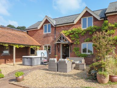 A patio with outdoor seating and a hot tub in front of a brick house with plants at Sherway Shippen in Clyst Hydon near Bradninch