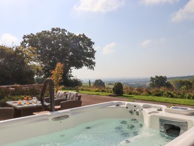 An outdoor hot tub with water jets near a table with chairs and drinks overlooking a countryside landscape at Sherway Shippen in Clyst Hydon near Bradninch