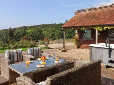 An outdoor patio with wicker chairs and a table set with croissants and juice near a wooden cabin and hot tub at Sherway Shippen in Clyst Hydon near Bradninch