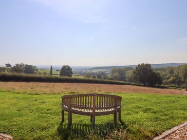 A wooden curved bench on grass facing fields and trees at Sherway Shippen in Clyst Hydon near Bradninch