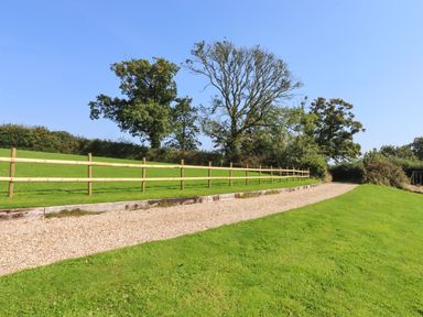 A gravel path next to a wooden fence and green grass with trees in the background at Sherway Shippen in Clyst Hydon near Bradninch
