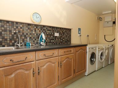A laundry room with wooden cabinets washing machines an iron a sink and a wall clock at Sherway Shippen in Clyst Hydon near Bradninch
