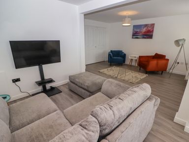 A living room with a grey sofa and ottoman a TV on a stand two armchairs a rug a side table a standing lamp and a painting on the wall at 69 Ffordd Y Meillion in Llanelli
