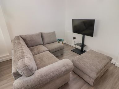 A small living room with a grey sectional sofa ottoman a TV on a stand and a small round side table at 69 Ffordd Y Meillion in Llanelli