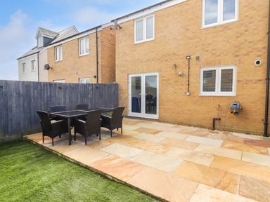 A backyard with a patio, a dining table with six chairs, a grass lawn, and a brick house with double doors at 69 Ffordd Y Meillion in Llanelli