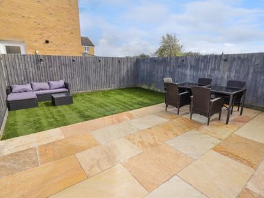 A backyard with a stone patio a grass area a sectional sofa and a dining table with chairs at 69 Ffordd Y Meillion Llanelli