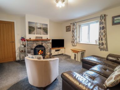 A living room with a stone fireplace a TV a brown leather sofa a white armchair and a window with curtains at Ysgubor Newydd Parc Isaf in Dyffryn Ardudwy