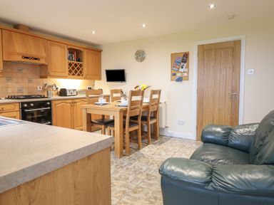 A kitchen and dining area with wooden cabinets and table next to a green leather armchair at Ysgubor Newydd Parc Isaf in Dyffryn Ardudwy
