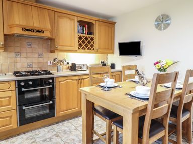 A kitchen with wooden cabinets a gas stove oven a dining table set with dishes and a wall clock at Ysgubor Newydd Parc Isaf in Dyffryn Ardudwy