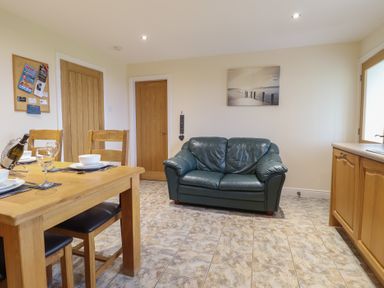 A dining area with a wooden table and chairs set with dishes and a bottle of wine a green leather sofa two wooden doors and wooden kitchen cabinets at Ysgubor Newydd Parc Isaf Dyffryn Ardudwy