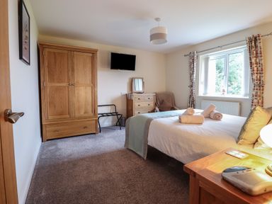 A bedroom with a bed with rolled towels a wooden wardrobe a chest of drawers a chair a wall-mounted television and a window with curtains at Ysgubor Newydd Parc Isaf in Dyffryn Ardudwy