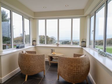 A seating area with two wicker chairs and a round wicker table with a tray and teapot by large windows overlooking a landscape at Ysgubor Newydd Parc Isaf in Dyffryn Ardudwy