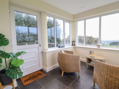 A sunroom with wicker chairs a small round wicker table a white door large windows and a potted plant at Ysgubor Newydd Parc Isaf in Dyffryn Ardudwy