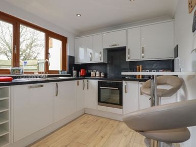 A kitchen with white cabinets a black countertop a built in oven a sink under a window and beige bar stools at 3 The Bowling Green in St Just in Roseland near St Mawes