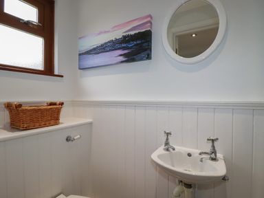 A bathroom with a toilet, a small sink, a round mirror, a window, a wicker basket, and a landscape picture on the wall at 3 The Bowling Green St Just in Roseland near St Mawes