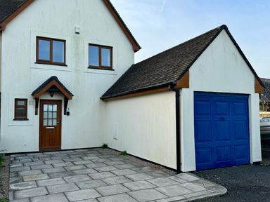 A two-story house with white walls brown framed windows a brown front door and a blue garage door at 3 The Bowling Green in St Just in Roseland near St Mawes