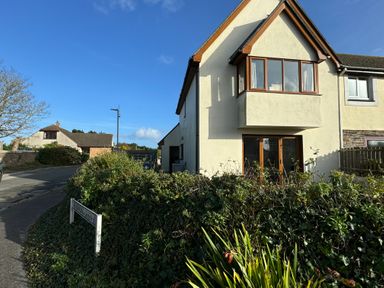 A residential street with a two-story house white walls and brown window frames surrounded by plants and bushes at 3 The Bowling Green in St Just in Roseland near St Mawes