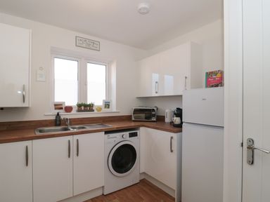 A kitchen with white cabinets a washing machine a sink a refrigerator and small plants on the windowsill at Cotswold Outlet Coach House in Tewkesbury