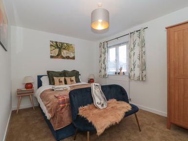 A bedroom with a double bed green and animal print pillows bedside tables a window with patterned curtains a wooden wardrobe and a blue bench with a fur throw and an owl cushion at Cotswold Outlet Coach House in Tewkesbury