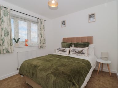 A bedroom with a double bed with green pillows and blanket two framed pictures above the bed window with patterned curtains and a small round side table with a lamp at Cotswold Outlet Coach House in Tewkesbury