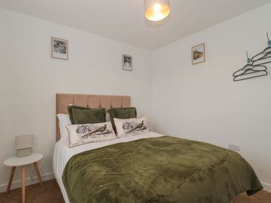 A bedroom with a bed with green pillows and blanket a white bedside table with a lamp three framed pictures on the wall and coat hangers on hooks at Cotswold Outlet Coach House in Tewkesbury