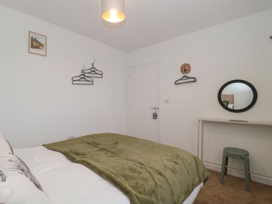 A bedroom with a bed covered with a green blanket a wall with hangers a round mirror above a simple desk and a stool at Cotswold Outlet Coach House Tewkesbury