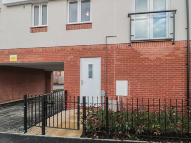 Exterior view of a building with red brick lower wall and white upper wall a white door with a small window a black metal gate and fence with plants behind it and a balcony above the door at Cotswold Outlet Coach House in Tewkesbury