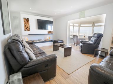 A living room with black leather sofas, a wall-mounted TV, a coffee table, and a window seating area at The Gables in Tywyn