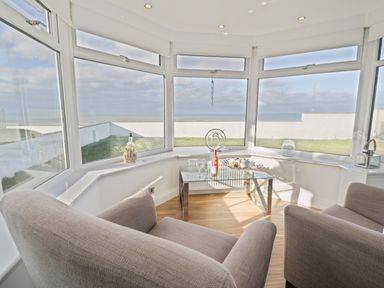 A sunroom with large windows overlooking a lawn and sea with two armchairs and a glass coffee table at The Gables in Tywyn
