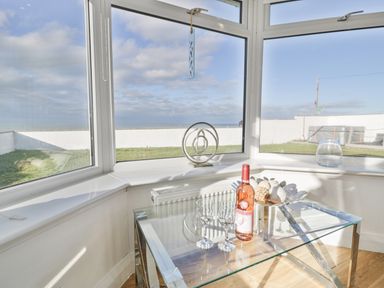 A glass table with wine bottle and glasses in a room with large windows overlooking a garden at The Gables in Tywyn