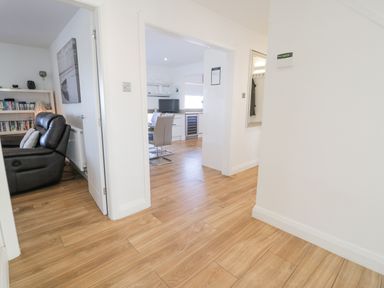 A wooden floor hallway leading to a living room with a black leather sofa and a kitchen with dining chairs at The Gables in Tywyn