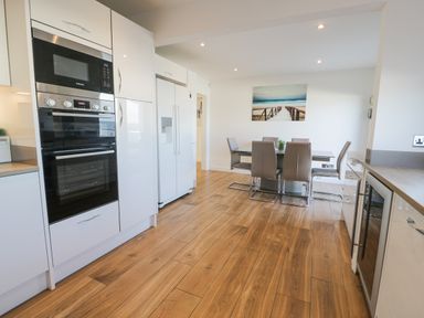 A kitchen and dining area with white cabinets wooden floor a dining table with six chairs and a wall painting at The Gables in Tywyn