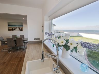 A kitchen sink with a vase of flowers on the windowsill overlooking a garden and sea with a dining area in the background at The Gables in Tywyn