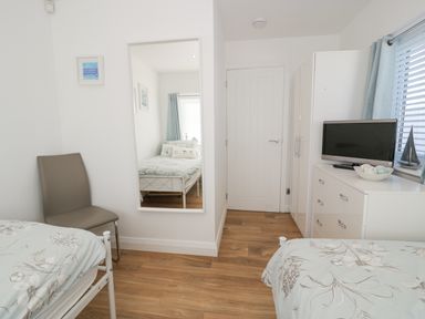 A bedroom with two beds a chair a wall mirror a white dresser with a television and a window with blinds and curtains at The Gables in Tywyn