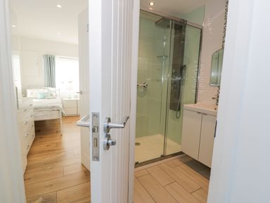 A bathroom with a glass shower enclosure and a white sink cabinet next to a bedroom with a bed and wooden floor at The Gables in Tywyn